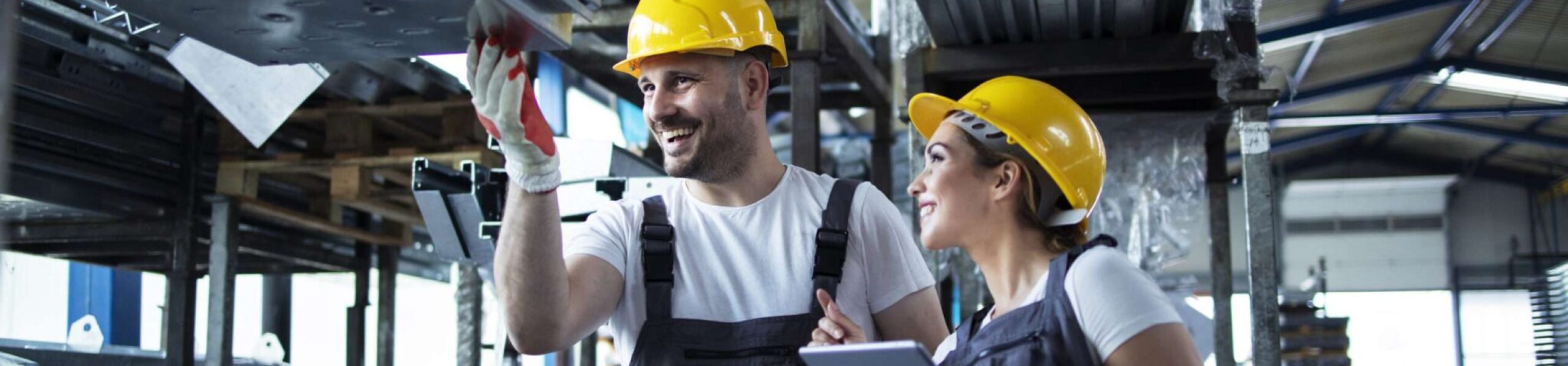 Factory workers checking inventory with tablet computer in industrial warehouse full of metal parts.