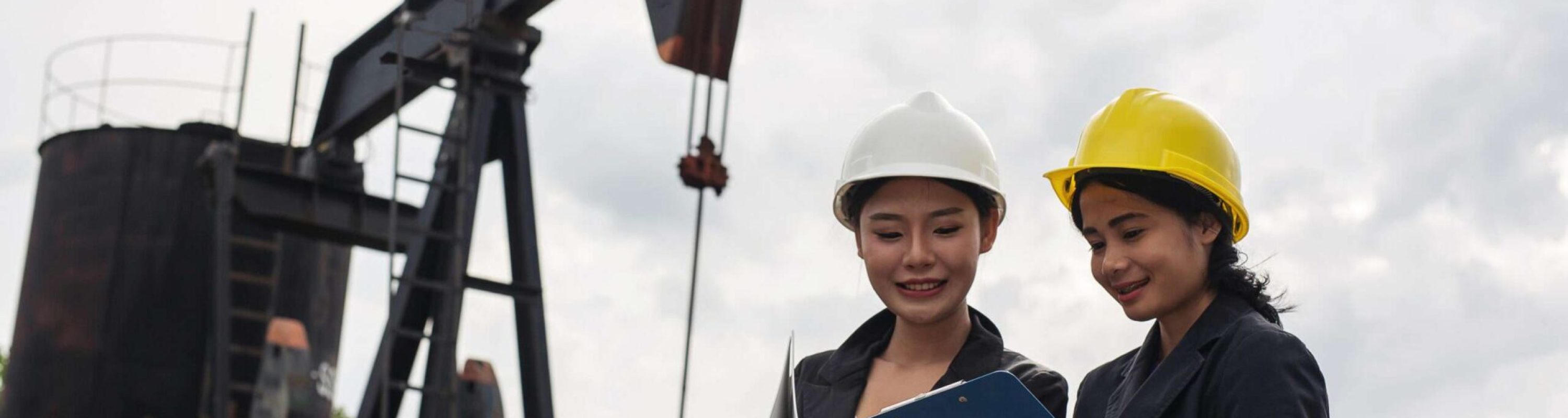 Two female engineers stand beside working oil pumps with a white sky background.